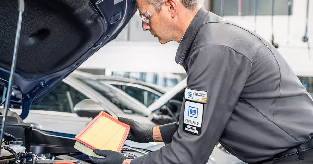 GM certified technician checking engine air filter in service center, wearing dark gray uniform with GM badge